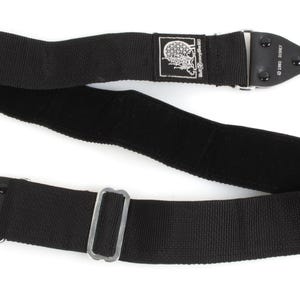 Peut inclure: Sangle d'appareil photo noire avec des clips métalliques et un repose-épaules rembourré. La sangle a une texture tissée et un petit patch avec un motif de crâne. La sangle est conçue pour être fixée à un appareil photo.