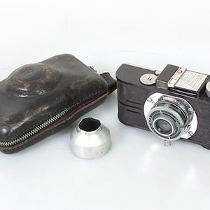 May include: A vintage black and silver camera with a leather case and a lens cap. The camera has a silver lens and a black body with silver trim.