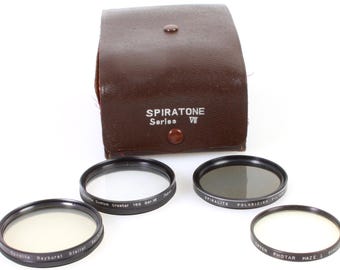 May include: A set of four camera lens filters in a brown leather case. The filters are labeled "Suralite Rayburst Grallar", "Siste Crostar 150 Ser W", "SPIRALITE POLARIZING FILTER", and "TOPEN PHOTAR HAZE 1", and are part of the "SPIRATONE Series VII" collection.
