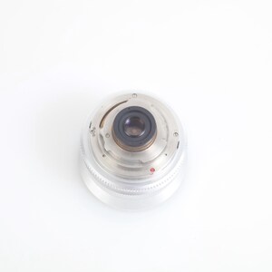 Pu&ograve; includere: Un obiettivo per fotocamera trasparente e circolare con un centro nero e componenti metallici argentati. L'obiettivo ha una copertura in plastica trasparente e scanalata ed &egrave; posto su uno sfondo bianco. Accessorio fotografico.