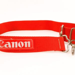 May include: A bright red camera strap made of woven nylon fabric. The strap features a rectangular patch with the white text Canon printed on it. It includes silver-colored metal hardware, including a sliding adjustment buckle and a swivel hook.