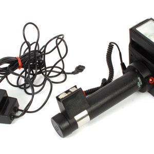 Puede incluir: Un flash Ascor light negro con un cargador de CA incorporado y un cable en espiral. El flash tiene un cuerpo de plástico negro con un anillo de metal plateado alrededor de la lente. El cargador de CA es una caja negra con una etiqueta blanca que dice "Ascor light Dual Charger AC Adapter".
