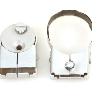 May include: Two silver-colored metal clamps with adjustable features. Each clamp has a circular top and a rectangular base with a split design. The clamps are designed to hold cylindrical objects.
