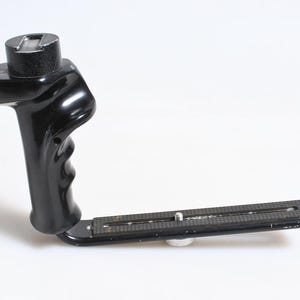 Puede incluir: Un agarre de cámara negro con un asa y una placa de montaje. El agarre tiene un asa texturizada para un agarre seguro y una montura superior para conectar una cámara. La placa de montaje tiene un tornillo para asegurar la cámara.
