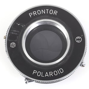 May include: A vintage Polaroid camera lens with the word "PRONTOR" at the top and "POLAROID" at the bottom. The lens is black with a silver rim and a circular aperture. The aperture blades are visible, and the lens is set against a white background.