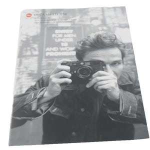 May include: Black and white photograph of a person holding a Leica M6 TTL 0.58 camera. The person is looking through the camera lens. The image has text at the top that reads "LEICA M6 TTL 0.58". The background is blurred.