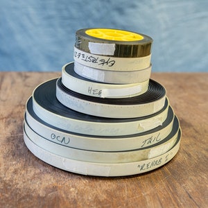 May include: A stack of six film reels, some with labels, on a wooden surface. The labels include "HEG", "OCN", "TRAIL", and "REHAB 2".