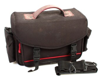 May include: Black camera bag with red accents and a detachable shoulder strap. The bag has a top handle and two adjustable buckles on the front.