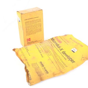 Pode incluir: Um pacote e uma caixa de revelador Kodak Microdol-X amarelos. O pacote está rotulado com o nome do produto e outras informações. A caixa é de cor castanho claro e também tem informações sobre o produto.