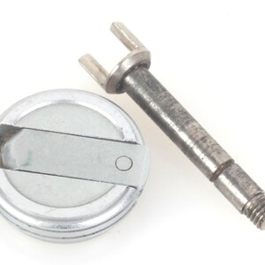 Puede incluir: Dos componentes mecánicos metálicos sobre fondo blanco. Uno es una pieza circular con un diseño ranurado y un pasador central. El otro es una varilla con un extremo bifurcado y una base roscada.