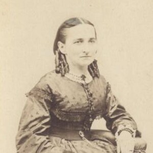 May include: A black and white portrait of a woman wearing a dark dress with lace trim. Her hair is styled in a braid with curls. She is looking directly at the camera.