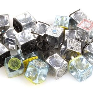 Op de afbeelding: Een verzameling vintage flashcubes, elk met een helder, kubusvormig ontwerp en een reflecterend interieur. Sommige kubussen hebben zwarte bases, terwijl andere logo's en tekst in verschillende kleuren hebben, waaronder geel, blauw en rood. De flashcubes liggen verspreid op een wit oppervlak.