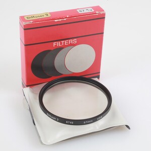 Puede incluir: Una caja roja con la etiqueta "FILTROS" con un juego de filtros negro y plateado. Un filtro difusor de 67 mm está en una funda de plástico transparente. El filtro tiene un borde negro.