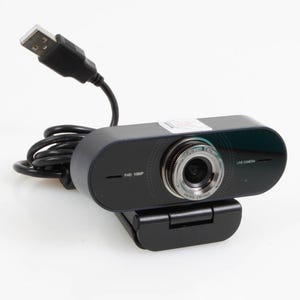 Puede incluir: Una webcam negra con lente plateada y cable USB. La webcam tiene el texto "FHD 1080P" y "LIVE CAMERA" impreso. La webcam está montada en un soporte negro ajustable.