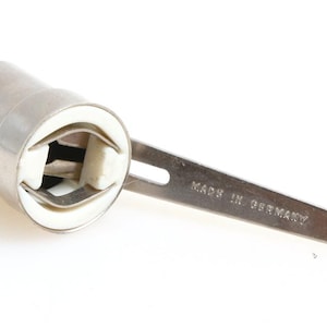 Puede incluir: Un portalámparas de automóvil plateado con interior de cerámica blanca y mango metálico. El mango tiene un agujero y el texto "MADE IN GERMANY".