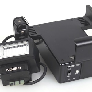 May include: A black Nissin camera flash unit with a rectangular head and a separate battery pack base. The base features a power switch labeled ON and OFF, a white test button, and the text Nissin FSP Made in Japan printed on the front.