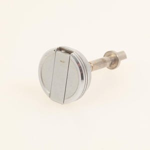 Puede incluir: Un pomo de metal plateado con una cara circular que presenta una ranura vertical central y un borde exterior elevado. El pomo está unido a un vástago metálico cilíndrico que se extiende hacia afuera, terminando en una pequeña base circular.