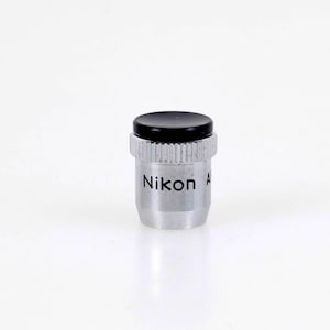 May include: A silver-colored Nikon microscope objective lens with a black circular cap on top. The cylindrical metal body features a knurled grip section near the top and the brand name Nikon printed in black lettering on the side of the barrel.