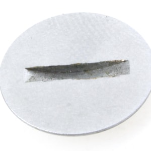 Puede incluir: Un objeto metálico circular de color plateado con una ranura rectangular cortada en su superficie. La ranura es de color gris oscuro y parece ser un hueco en el metal. El objeto está sobre un fondo blanco.