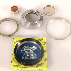 May include: An Ednalite precision filter holder set including a black circular filter holder, a yellow cardboard box with the text Ednalite Precision Filter Holder, two small circular glass filters, and three silver-toned metal adapter rings.