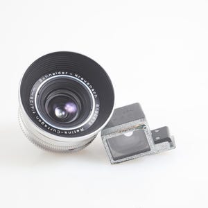 Pu&ograve; includere: Un obiettivo per fotocamera vintage con finitura nera e argento, etichettato "Schneider-Kreuznach Retina-Curtagon f/4 28mm". Un mirino rettangolare &egrave; posizionato a destra dell'obiettivo. Gli elementi sono su sfondo bianco.