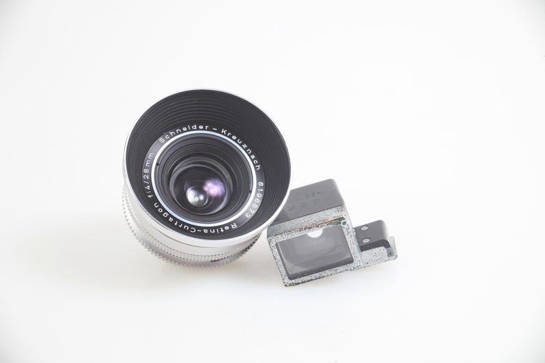 Pu&ograve; includere: Un obiettivo per fotocamera vintage con finitura nera e argento, etichettato "Schneider-Kreuznach Retina-Curtagon f/4 28mm". Un mirino rettangolare &egrave; posizionato a destra dell'obiettivo. Gli elementi sono su sfondo bianco.