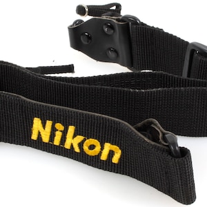 May include: Black Nikon camera strap with yellow embroidered Nikon logo. The strap is made of a durable woven material and features adjustable buckles and leather accents. Ideal for DSLR cameras.