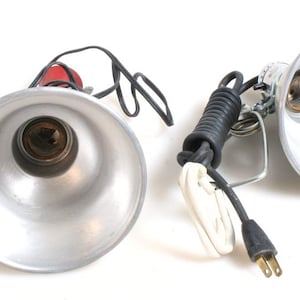 Peut inclure: Deux lampes de travail en métal argenté avec des cordons et des fiches noirs. Les lampes ont une douille en céramique ronde blanche et un abat-jour en métal argenté.