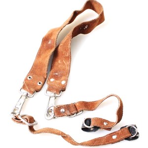 May include: Brown leather camera strap with metal clasps and connectors. The strap is made of worn, distressed leather and features a loop for attaching to a camera. The strap is designed to be worn around the neck or shoulder.
