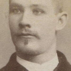 May include: Sepia-toned portrait of a man with light hair and a mustache. He wears a dark jacket over a white collared shirt. The photograph is a close-up of his face and upper body.