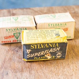 Puede incluir: Tres cajas vintage de bombillas flash Sylvania Superflash. Las cajas son amarillas y negras con detalles verdes y blancos. El texto en las cajas dice "Sylvania Superflash Press 40 Flashbulb".