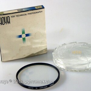 Puede incluir: Un filtro difusor para cámara Hoya con borde de metal negro, lente de cristal circular transparente y caja de cartón blanco a juego con el logotipo de Hoya, un gráfico de cruz azul y verde y el texto FOR TECHNICAL PHOTOGRAPHY.