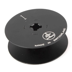 Puede incluir: Un carrete de película de plástico negro con una etiqueta blanca que dice "Bolex Switzerland" y "Made in Switzerland". El carrete tiene un agujero central y un número "1" impreso en la parte superior.