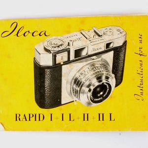 Puede incluir: Un manual de instrucciones amarillo para la cámara Iloca Rapid I+IL+II-IIL. La portada presenta una cámara negra y plateada con el nombre de la marca "Iloca" impreso en cursiva.