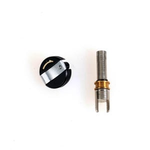 May include: A black and silver circular control knob featuring a small gold arrow indicator alongside a separate cylindrical metal shaft component with a gold-colored ring and a slotted base, both displayed against a plain white background.