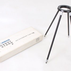 May include: A black Canon Handy Stand F tripod with extension tube M5, in its original box. The tripod has three legs and a round top with a screw mount.