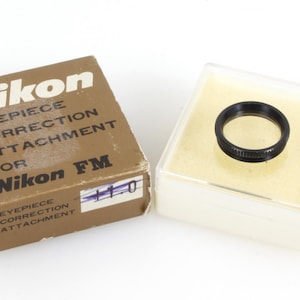 Peut inclure: Un adaptateur de correction d'oculaire Nikon pour Nikon FM, avec une bague noire dans un étui en plastique transparent et une boîte marron. La boîte porte le logo et le texte Nikon. L'accessoire est pour un appareil photo.