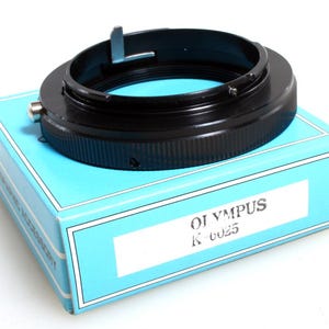 Puede incluir: Un anillo adaptador de objetivo de cámara Olympus K-6025 negro descansa sobre una caja azul claro con ribete blanco. El anillo adaptador es circular con un exterior estriado. La caja tiene el texto "OLYMPUS K-6025" impreso.
