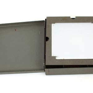 May include: A grey metal box with a hinged lid. The box has a white rectangular insert inside. The box is open and the lid is propped up.