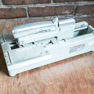 May include: A silver and gray Singer industrial thread cutter with a handle and a ruler on the top. The cutter has a label that reads "Singer Industrial Thread Cutter Model No. Serial No."