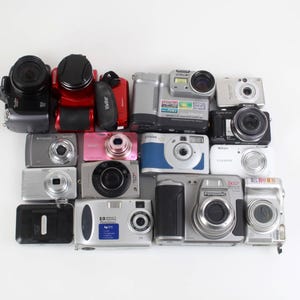 May include: A collection of various digital cameras arranged in a rectangular grid. The assortment includes multiple compact and bridge models in black, silver, red, pink, blue, and white finishes, featuring diverse lens sizes and body designs.