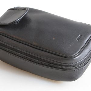 Può includere: Un astuccio in pelle nera con chiusura a zip e un lembo curvo sulla parte superiore. L'astuccio rettangolare presenta un piccolo logo in rilievo sul davanti. Probabilmente progettato per contenere piccoli oggetti.
