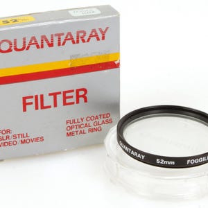 May include: A Quantaray 52mm Foggilizer filter for SLR, still, video, and movies. The filter has a black metal ring and clear optical glass. The box is grey with red and yellow stripes and the word FILTER in large red letters.