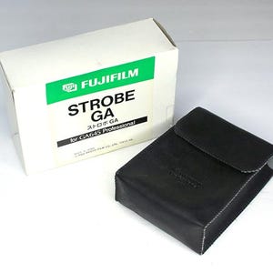 Op de afbeelding: Een witte doos met groene tekst die "FUJIFILM STROBE GA" en "for GA645 Professional" zegt. Een zwart leren etui is op de voorgrond.