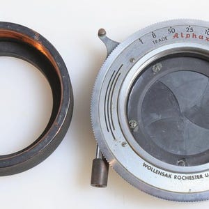 May include: A silver and black camera lens with a circular aperture and a dial that reads "Alphax" and "Trade Mark".