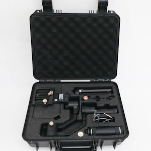 May include: Black hard-shell carrying case with an open lid, revealing a video camera stabilizer and accessories. The interior is lined with black foam cutouts to hold the equipment securely. The case has two black latches and a handle.