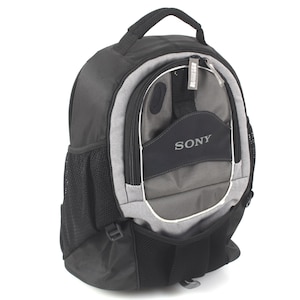 May include: A black and gray backpack featuring a top handle, a front zippered compartment with the Sony logo printed in white, and black mesh side pockets. The design includes a structured main body with multiple sections and silver zipper pulls.