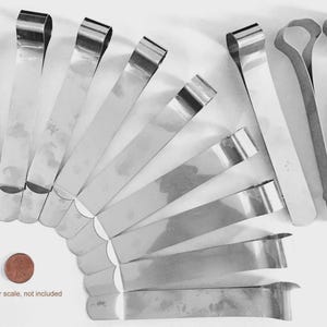 May include: A collection of twelve silver-coloured metal clips arranged in a fan shape. Each clip features a long, flat, rectangular body with a folded, rounded top end. A single copper-coloured penny is positioned near the bottom left for scale.