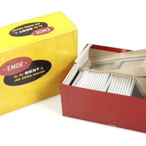 May include: A yellow box with the text "EMDE" and "for the BEST in slide binding materials" next to a red box filled with slide binding materials. The boxes are on a white background. The boxes are rectangular.