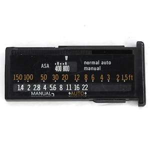 May include: Black camera light meter with white and orange text. The meter displays ASA, f-stops, and distance in feet. The text includes "normal auto manual" and "manual auto".